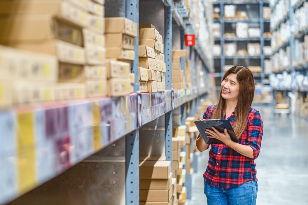 Asian woman warehouse worker standing and checking goods stock in warehouse and writing in clipboard for noted, shopping and self service, business furniture and storehouse industry concept