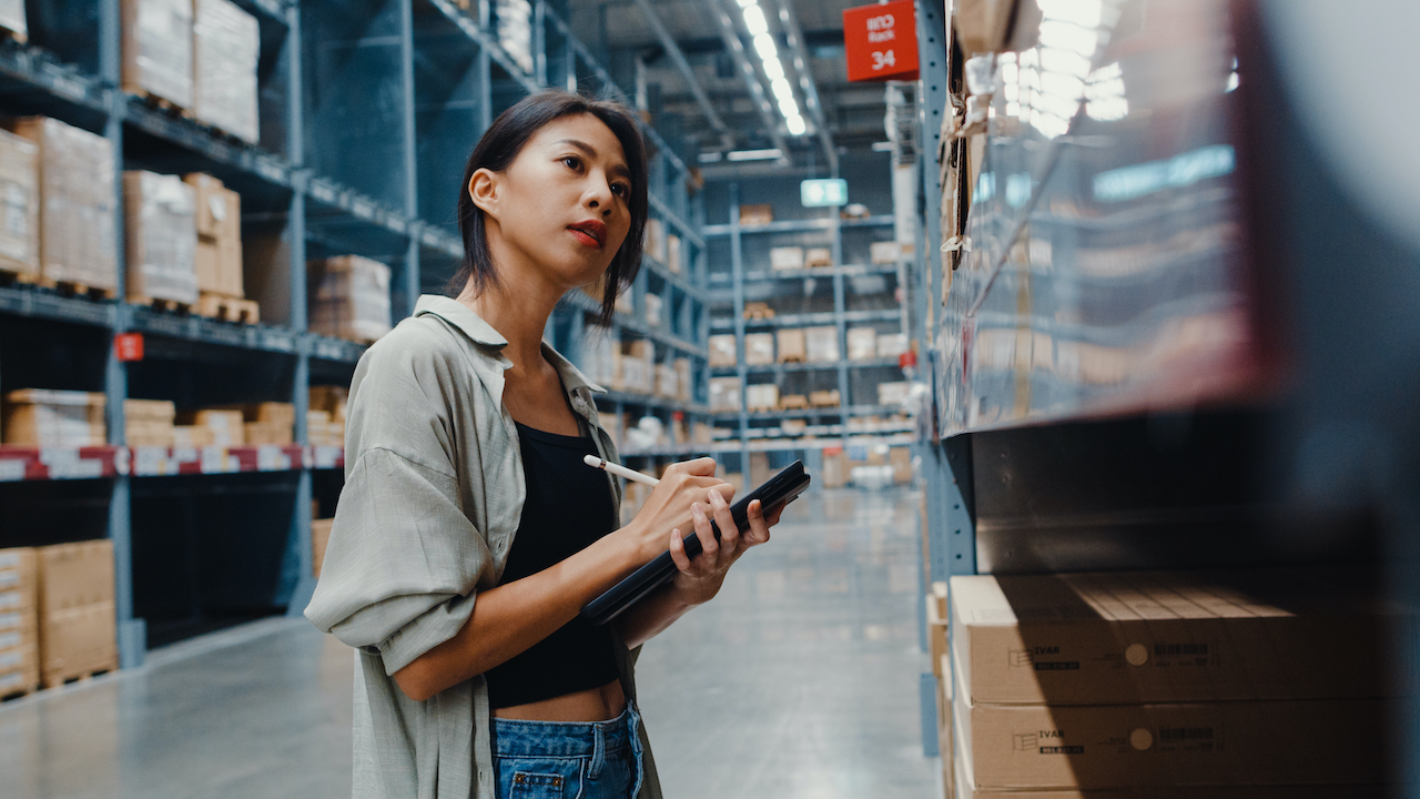 Attractive young Asia businesswoman manager looking for goods using digital tablet checking inventory levels standing in retail shopping center. Distribution, Logistics, Packages ready for shipment.