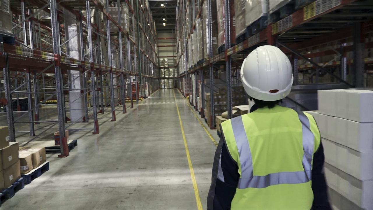 Back view portrait of warehouse manager and worker in hardhats doing stock inventory in warehouse, looking up at tall shelves with goods. Warehouse worker on hoverboard.