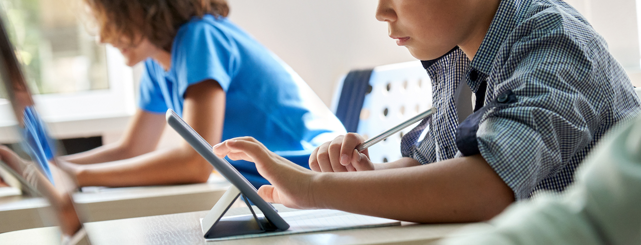 Close up cut shot of concentrated diligent Asian ethnicity elementary schoolboy touching using tablet device sitting in classroom with group of schoolchildren. Modern education technologies concept.