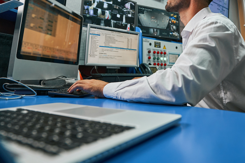 Caucasian system administrator in armchair looking at camera footage of server room on display