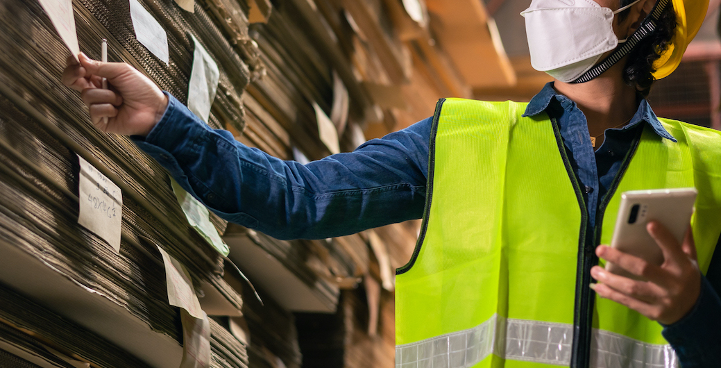 Engineer  wearing safety helmet and vest holding clipboard and take note on the paper .The working during outbreak of Covid-19 concept: Female warehouse staff with face mask use tablet check goods in warehouse.