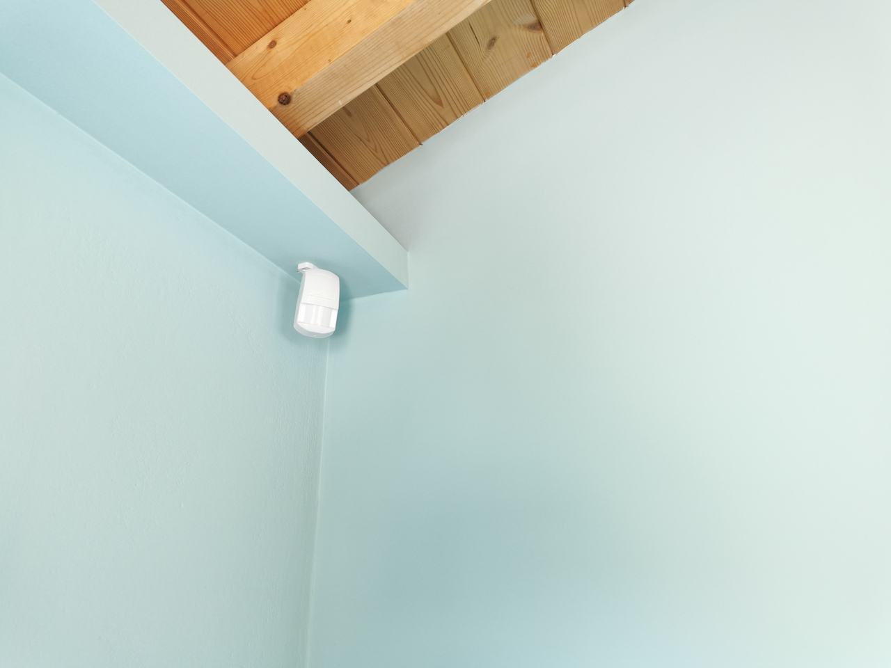 Motion sensor or detector for security system mounted on blue wall in mansard room with wooden ceiling.