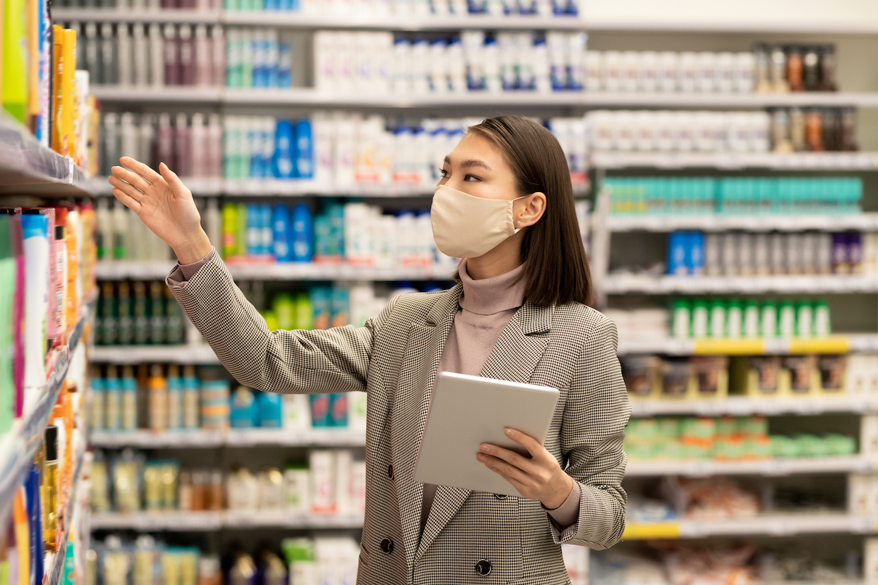 Young Asian female consumer with tablet standing by shelf with bodycare products and choosing one for herself while using touchpad