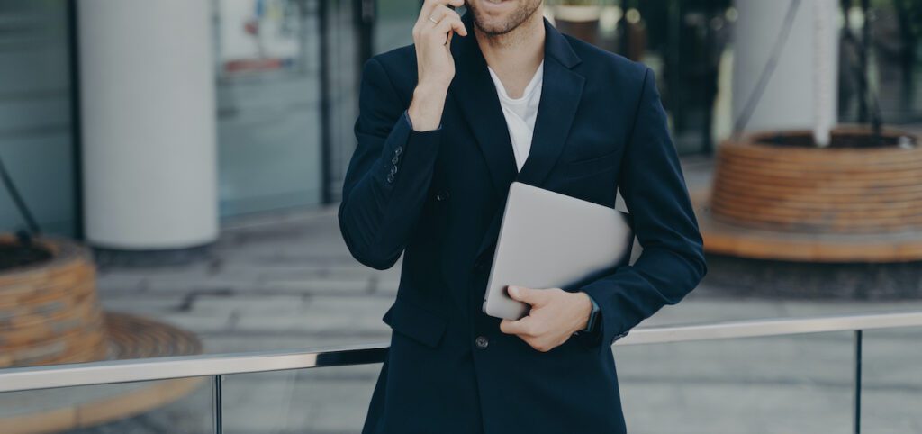 Half length shot of handsome businessman makes phone call in roaming using tariffs carries digital device looks away into distance wears formal clothes. Executive manager has mobile talk outdoors