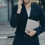 Half length shot of handsome businessman makes phone call in roaming using tariffs carries digital device looks away into distance wears formal clothes. Executive manager has mobile talk outdoors