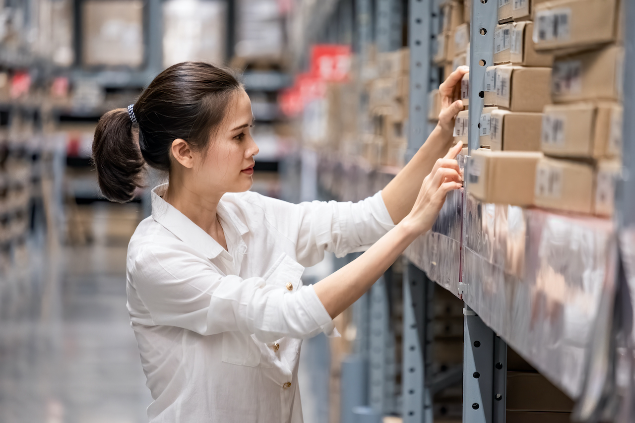 Warehouse Management System. Smart Asian young woman happy to work in store warehouse. Girl standing and checking quality and quantity of inventory stock on shelf by model name and serial number it show on packaging of goods.