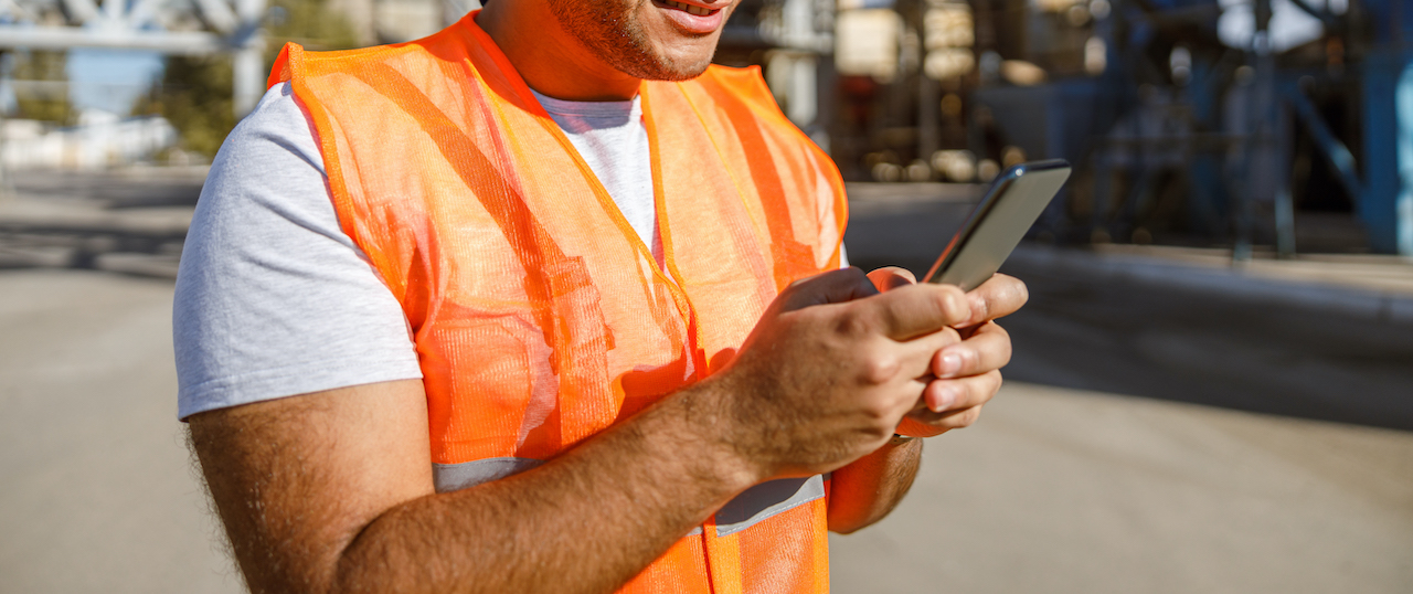 Concentrated man worker wearing blue cap and orange vest browsing cellphone at manufacturing facility of plant